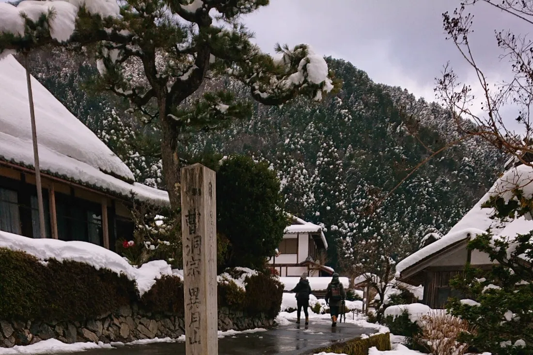 Snow on the thatched roofs along a lane in Miyama Kayabuki-no-Sato, with wooded hills behind